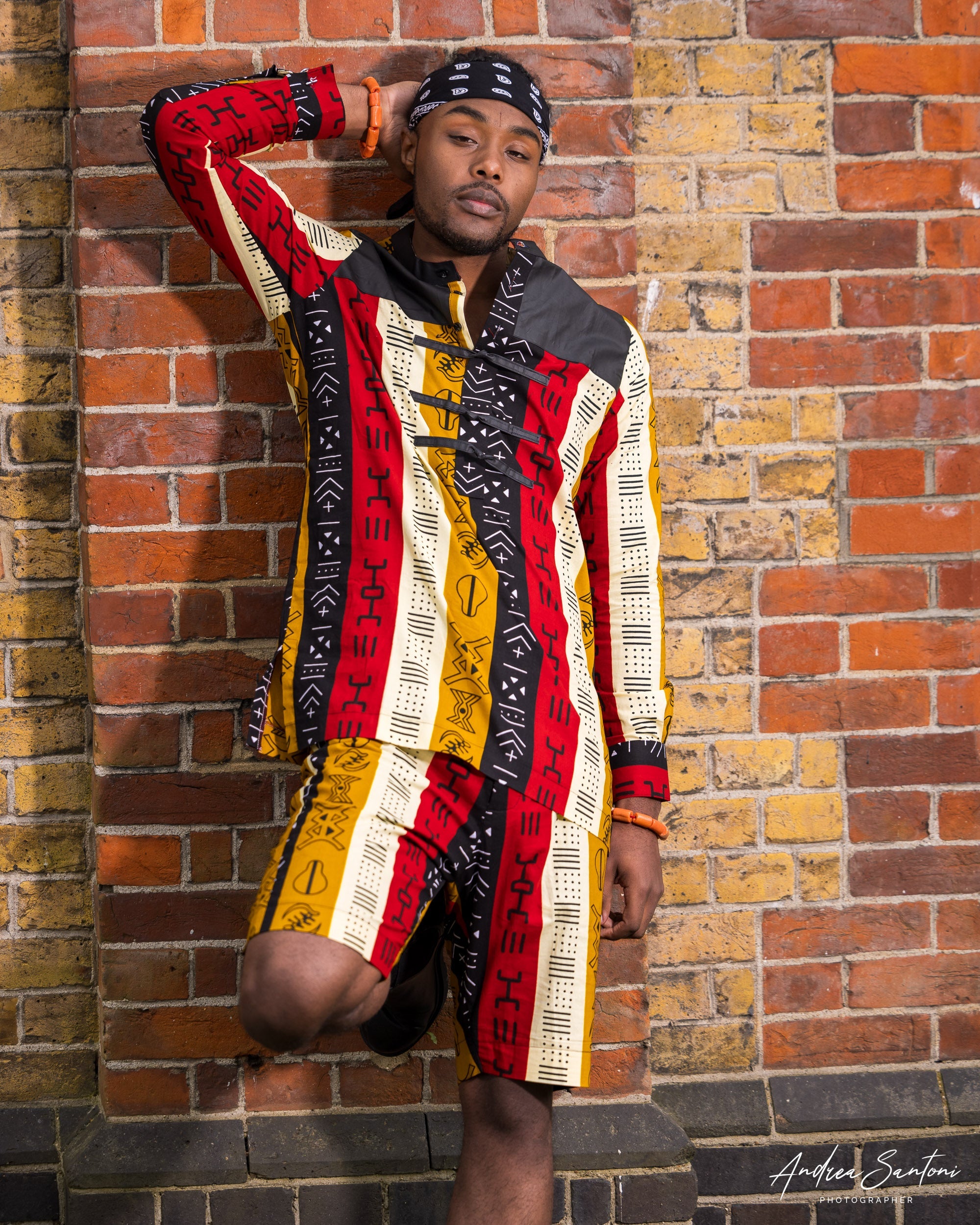 Male model wearing printed shirt and shorts standing and leaning against the wall with his hand behind his head