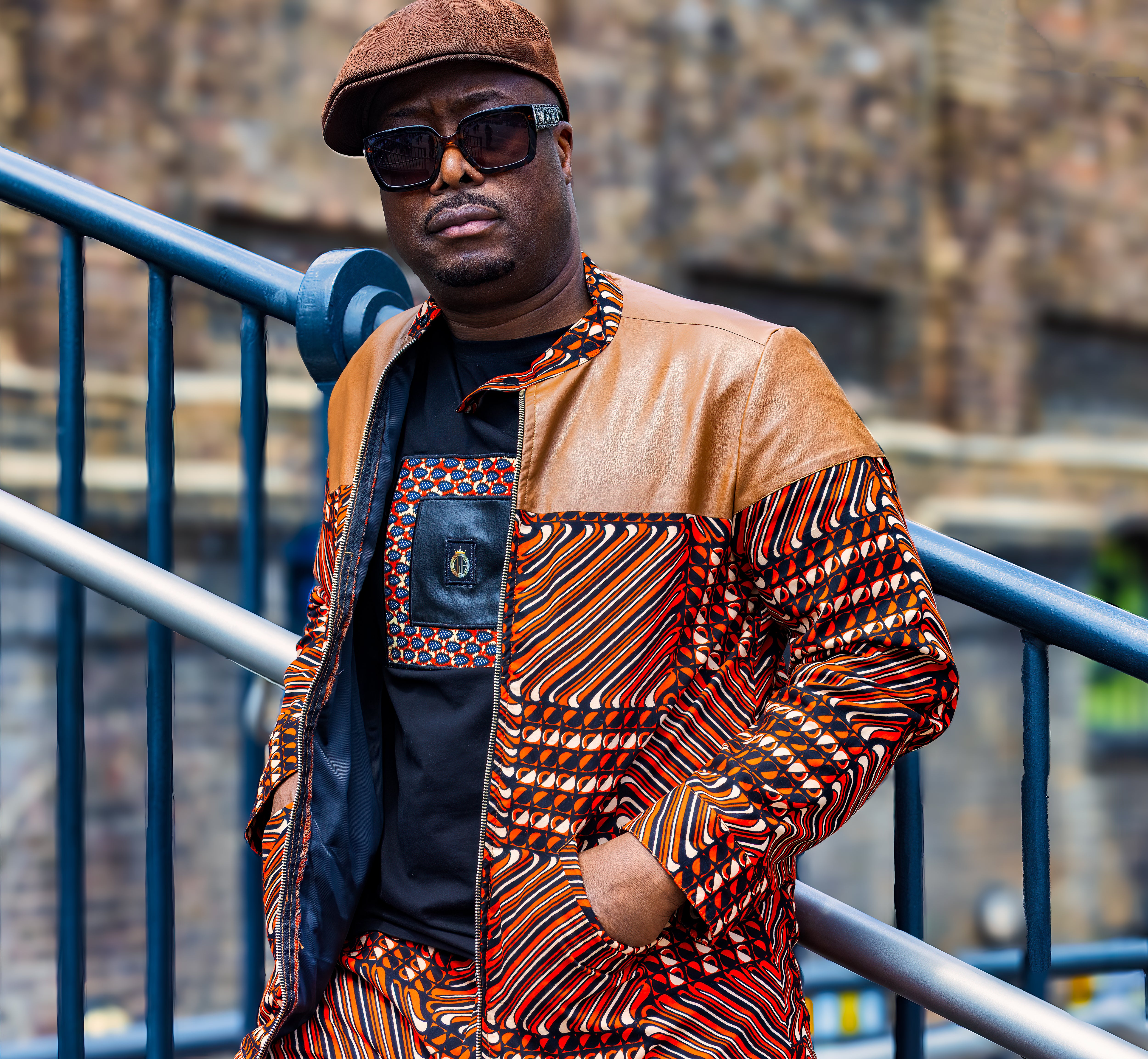 Man wearing a patterned Ankara print jacket and sunglasses standing against a metal railing.