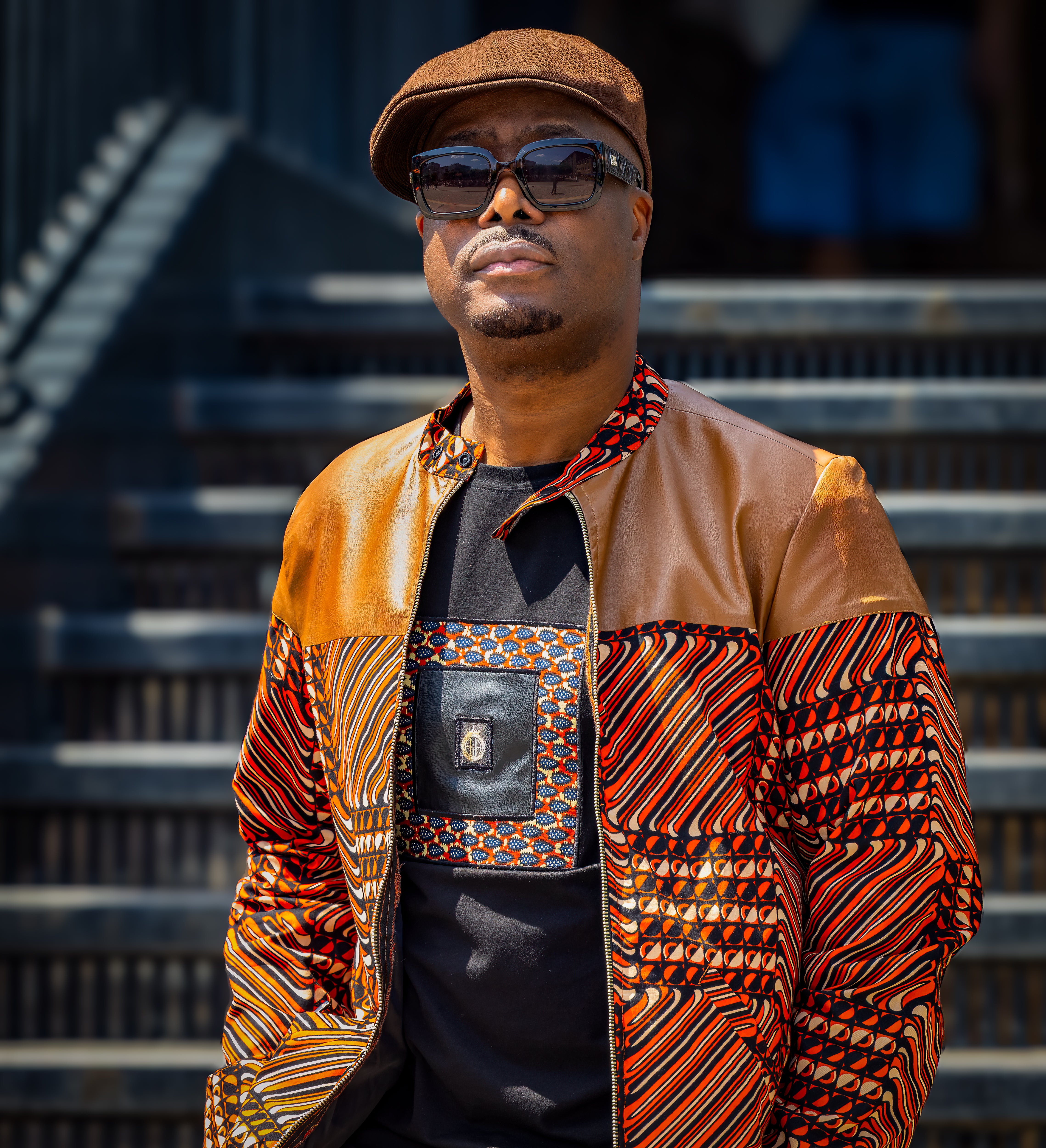 Man wearing African print logo t shirt and a patterned Ankara jacket and sunglasses standing on steps.